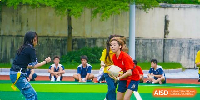 Students in various colored athletic shirts sit on the edge of an outdoor sports field during a physical education lesson at St Bees Dongguan.