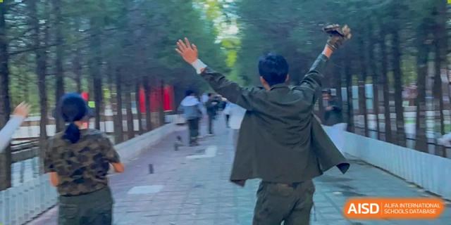 A student raises their arms in a joyful gesture while walking along a tree-lined campus path at Beijing No. 35 High School International Department.