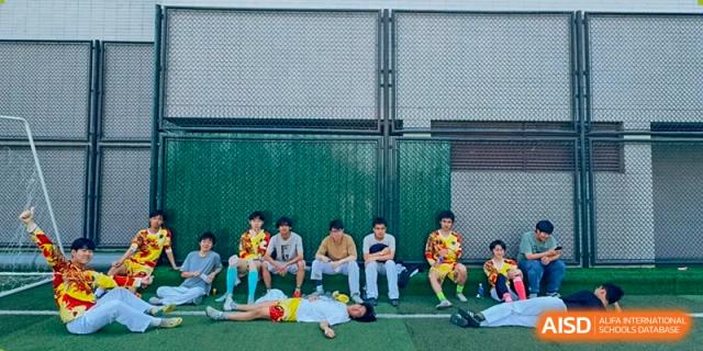 A group of students in sports jerseys rests on the green turf by a perimeter fence at Beijing No. 35 High School International Department.