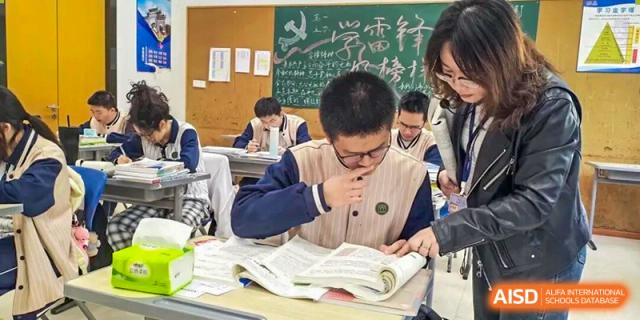 A focused high school student studies from an open textbook while a teacher assists nearby in a busy classroom at Xiamen ITG-SUIS High School