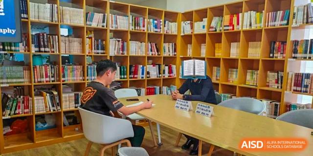 Students sit across a table reading and discussing academic materials in a quiet, well-organized library surrounded by wooden bookshelves at Xiamen ITG-SUIS High School