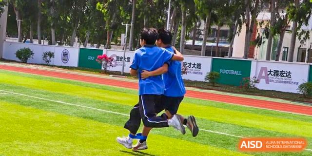 Students in blue sports uniforms supporting each other while running together on a green field, demonstrating teamwork, friendship, and cooperation during a school sports event at Xiamen ITG-SUIS High School