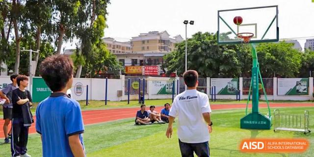 Students resting on the outdoor sports field while a teacher stands nearby supervising an activity, reflecting guided physical education, teamwork, and outdoor learning at Xiamen ITG-SUIS High School