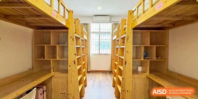 A well-lit student dormitory corridor featuring wooden bunk beds, storage shelves, and windows providing natural light in a clean residential space at Xiamen ITG-SUIS High School