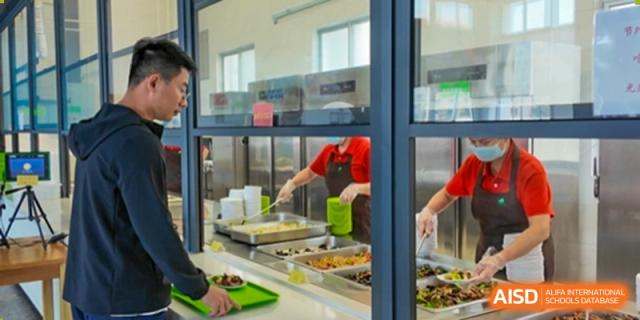 A cafeteria staff member serves food to a student using trays behind a glass counter, highlighting daily meal service and hygiene standards at Xiamen ITG-SUIS High School