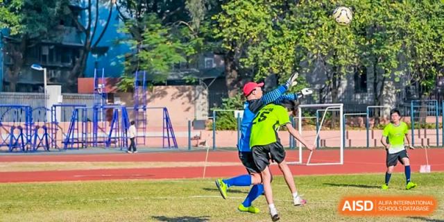 A goalkeeper in a blue jersey and red cap makes a high save during a soccer match on a sunny grass field, demonstrating athletic skill and teamwork at the Huafu International Department.