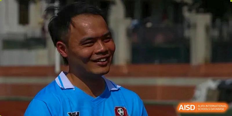 A close-up portrait of a smiling man wearing a blue athletic jersey with a soccer crest, captured during a bright outdoor sports competition at the Huafu International Department.