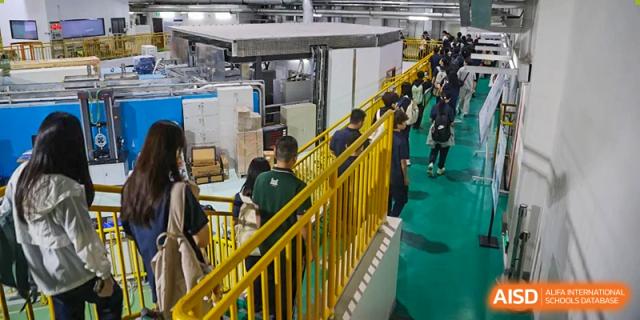 Students in green vests walking along a yellow-railed elevated walkway inside a large industrial or academic facility at the Huafu International Department.