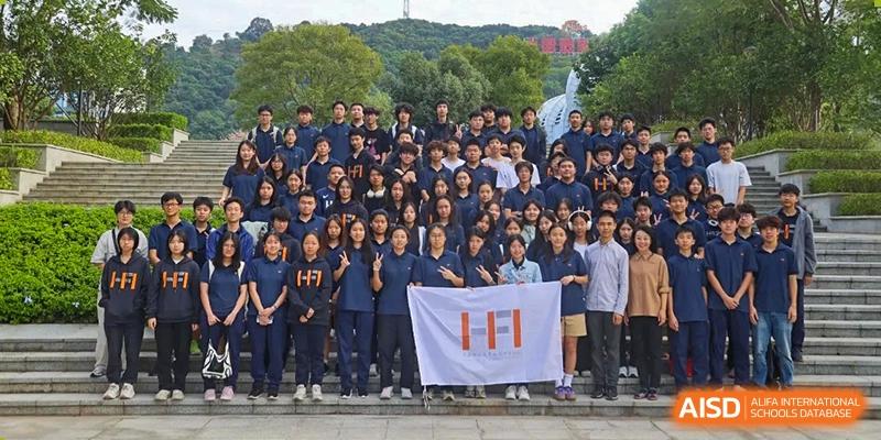 A tall vertical shot of a large student body gathered on stairs, holding an HFI logo banner during a formal event at the Huafu International Department.