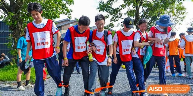 Three students participate in a three-legged race outdoors, wearing red and blue team vests while focusing on their synchronized steps during a sports day event at Huafu International Department.