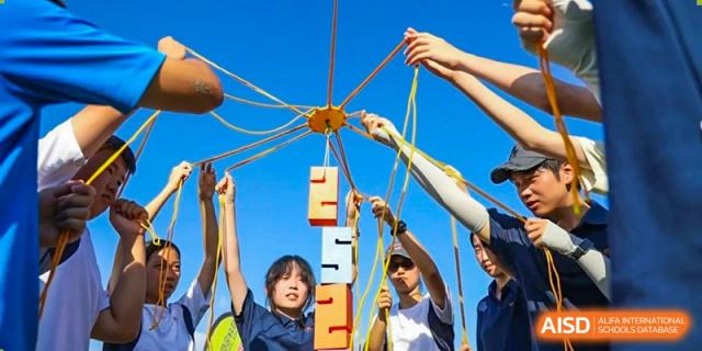 Students work together using strings to balance and stack large wooden blocks, practicing communication and teamwork during an outdoor leadership activity at Huafu International Department.