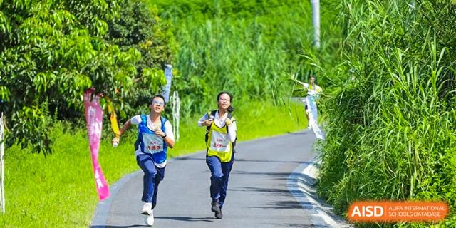 Two students jog together along a paved path surrounded by lush green hills, enjoying the fresh air and physical exercise during an organized school outing at Huafu International Department.