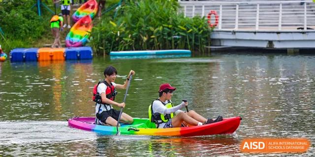 Two students navigate a vibrant, multi-colored kayak together, practicing their rowing technique during a sunny afternoon water sports lesson at Huafu International Department.