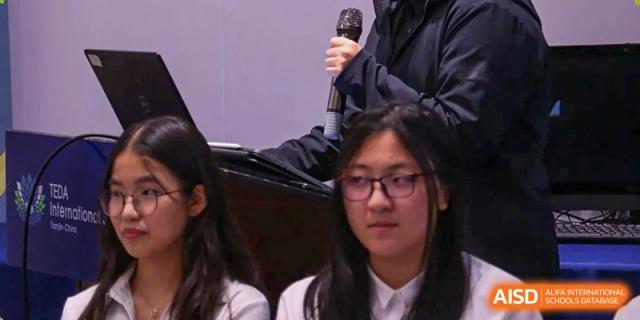 A student with glasses looks on attentively during a presentation or ceremony in a school hall at TEDA International School Tianjin.