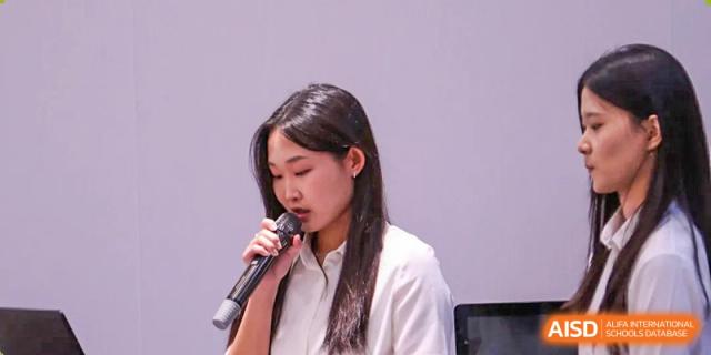 A young woman speaks into a microphone while reading from a paper during a National Honor Society induction at TEDA International School Tianjin.