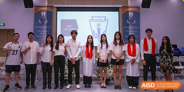 A group of students in white shirts and black pants stand on stage for a National Honor Society ceremony at TEDA International School Tianjin.