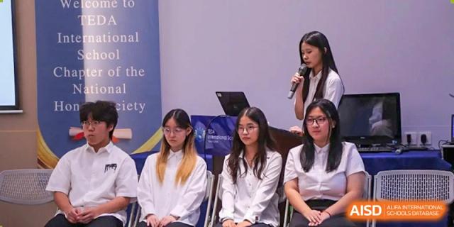 Students in white uniforms sit together during a school ceremony while a speaker presents from the podium at TEDA International School Tianjin.