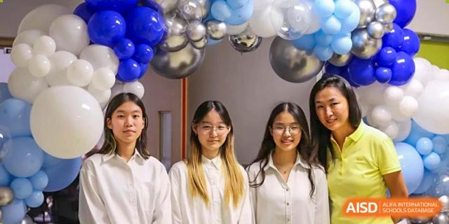 Three young female students in white shirts smile together in front of a festive blue and silver balloon arch at TEDA International School Tianjin.