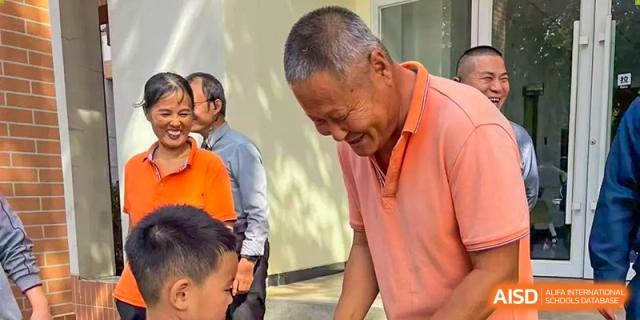 An older man in an orange shirt smiles warmly while interacting with younger students outdoors on campus at TEDA International School Tianjin.