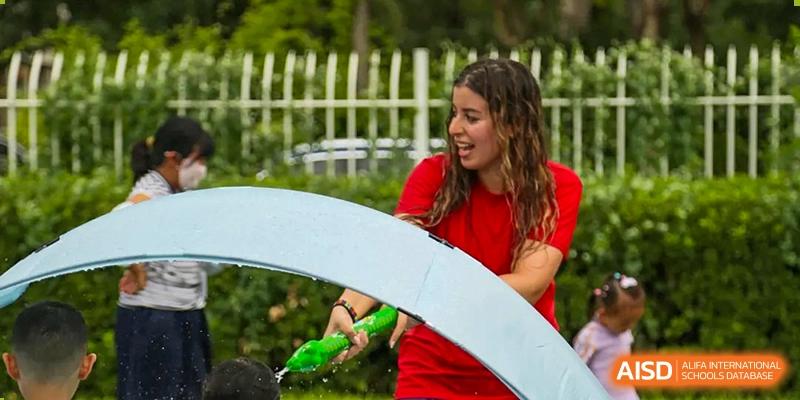 A teacher in a red shirt smiles while supervising children playing with water toys at TEDA International School Tianjin.