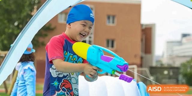 A boy in a blue swim cap laughs while aiming a large colorful water gun at TEDA International School Tianjin.
