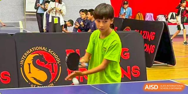 A student in a bright neon green shirt focuses on a table tennis match in the gymnasium at TEDA International School Tianjin.