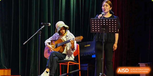 A student guitarist performs a solo piece while a teacher provides vocal accompaniment on stage at TEDA International School Tianjin.