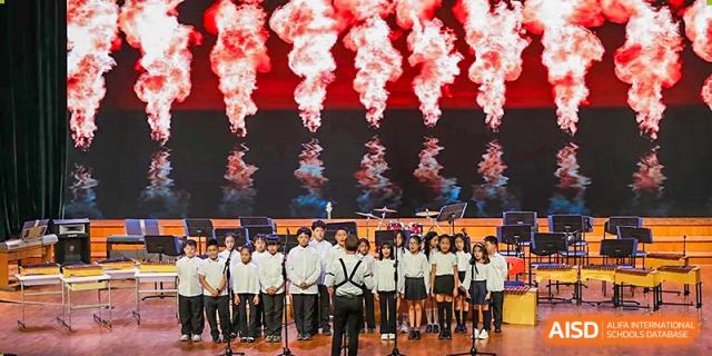 A large student choir performs together on stage in front of a dramatic red digital background at TEDA International School Tianjin.