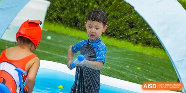 A young boy in a blue swim shirt has fun playing with a water sprayer during an outdoor activity at TEDA International School Tianjin.