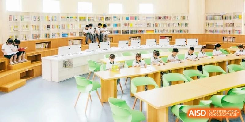 A bright, modern school library with students in uniform sitting at green chairs and light wood tables, reading books amidst shelves of resources at Ronghaui International School.