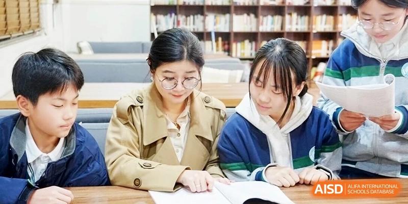 Two female students in casual attire sit together at a wooden table in a library studying a textbook, focusing on their academic work at Ronghaui International School.