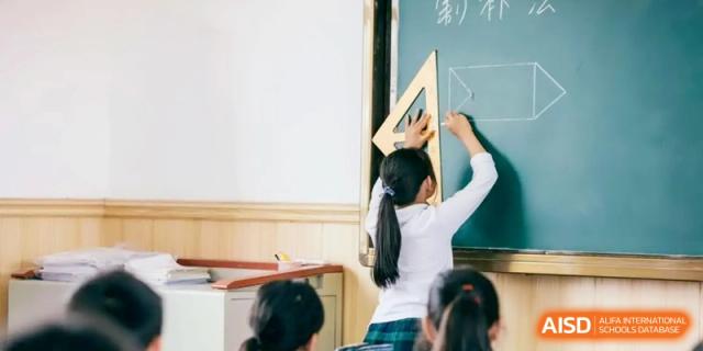 A student standing at the front of the classroom writing geometric shapes on a green chalkboard using a large wooden ruler and triangle at Ronghaui International School.