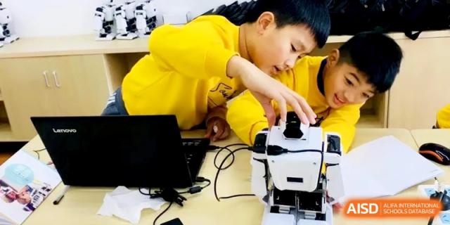 Two young students in yellow shirts working together to assemble and program a small white educational robot on a table, learning STEM skills at Ronghaui International School.