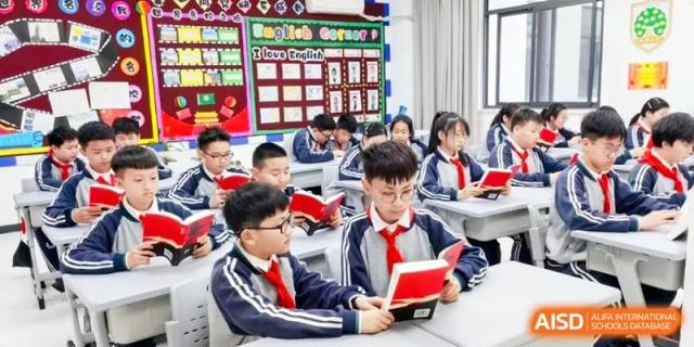 A lively classroom environment where young students in school uniforms are sitting at desks, reading books together in the English Corner area at Ronghaui International School.