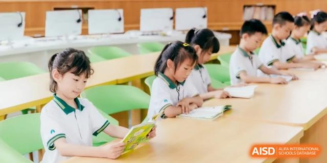 Young students in green and white uniforms sit at light green chairs, quietly reading books in a brightly lit school library at Ronghaui International School.