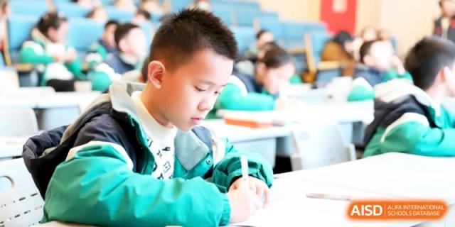 A young boy in a green jacket concentrates on writing in his notebook while seated at a desk in a classroom setting at Ronghaui International School.