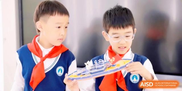 Two young students, wearing red scarves, examine a detailed model ship they have built together during a STEM class activity at Ronghaui International School.