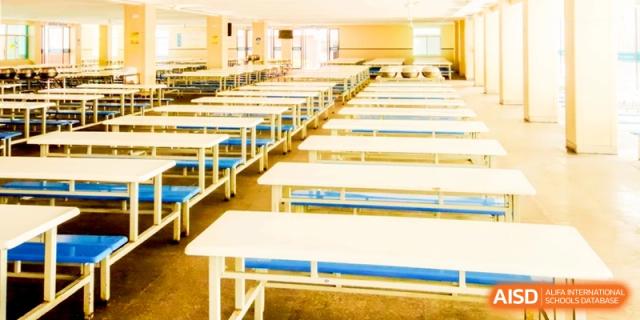 An empty school dining hall featuring rows of long white tables and blue chairs, prepared for lunch service for the student body at Ronghaui International School.