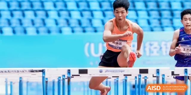 A male student athlete sprinting rapidly and jumping over a hurdle on a blue athletic track during a track and field meet at Ronghaui International School.