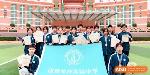 A group of students proudly holding up medals and certificates in front of the school building, celebrating a tournament victory at Ronghaui International School.