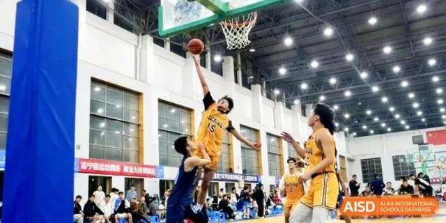 A male basketball player jumping high to shoot a ball into the hoop during an intense indoor game surrounded by teammates at Ronghaui International School.