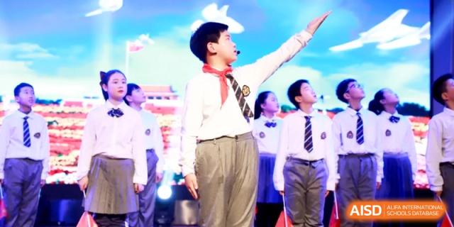 A student in a white uniform speaking passionately into a microphone on stage, leading an assembly with confidence at Ronghaui International School.