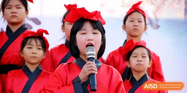 A young girl dressed in a traditional red outfit singing into a microphone on a stage during a school performance at Ronghaui International School.