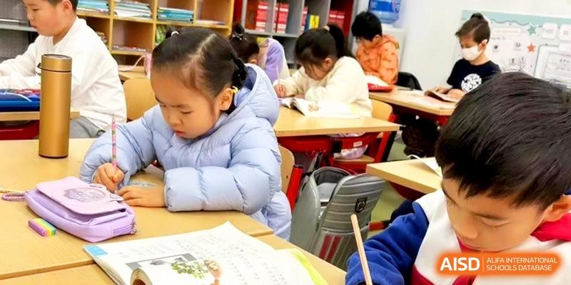 A young girl in a blue puffer jacket focuses intently on her classwork at a wooden desk at Shanghai Gold Apple Bilingual School.