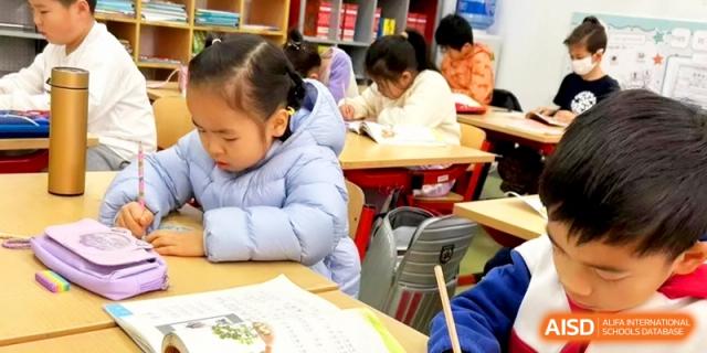 A young girl in a blue puffer jacket focuses intently on her classwork at a wooden desk at Shanghai Gold Apple Bilingual School.