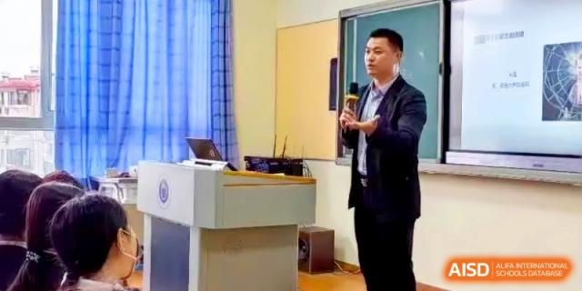 A male instructor in a suit speaks into a microphone while standing at a podium in a classroom at Shanghai Gold Apple Bilingual School.