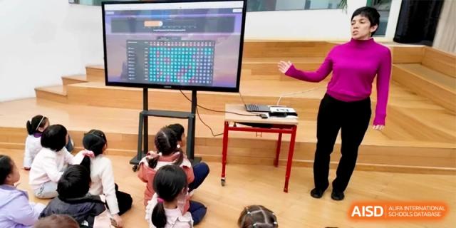 Students sit on the floor facing a large digital display used for a coding or math lesson at Shanghai Gold Apple Bilingual School.
