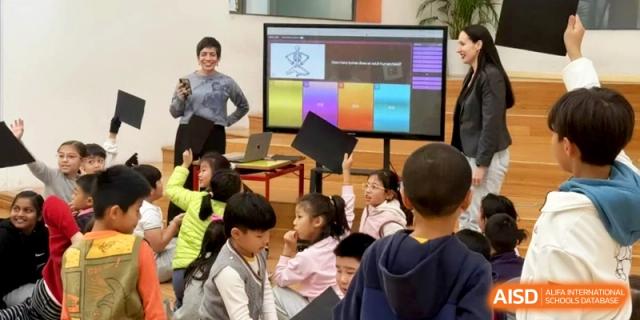 A teacher leads a group of students seated on the floor in front of a colorful digital screen at Shanghai Gold Apple Bilingual School.