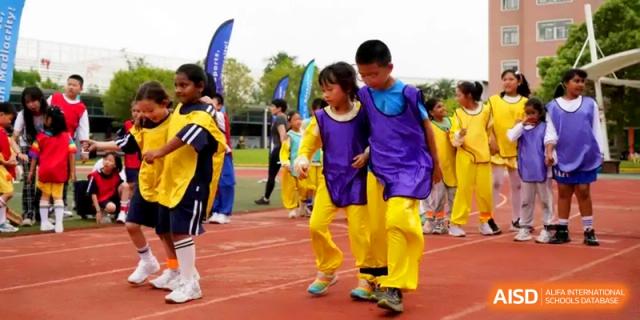 Two students wearing purple and yellow athletic bibs participating in a three-legged race at Shanghai Gold Apple Bilingual School.