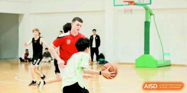 A student in a red shirt guards another student dribbling a basketball indoors at Shanghai Gold Apple Bilingual School.